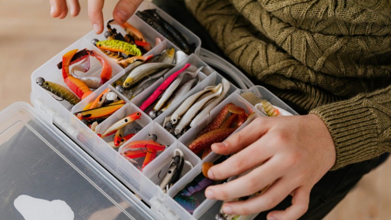 Close-up of assorted fishing lures neatly arranged in a plastic tackle box held by a person., tags: salt pencil der für - pexel